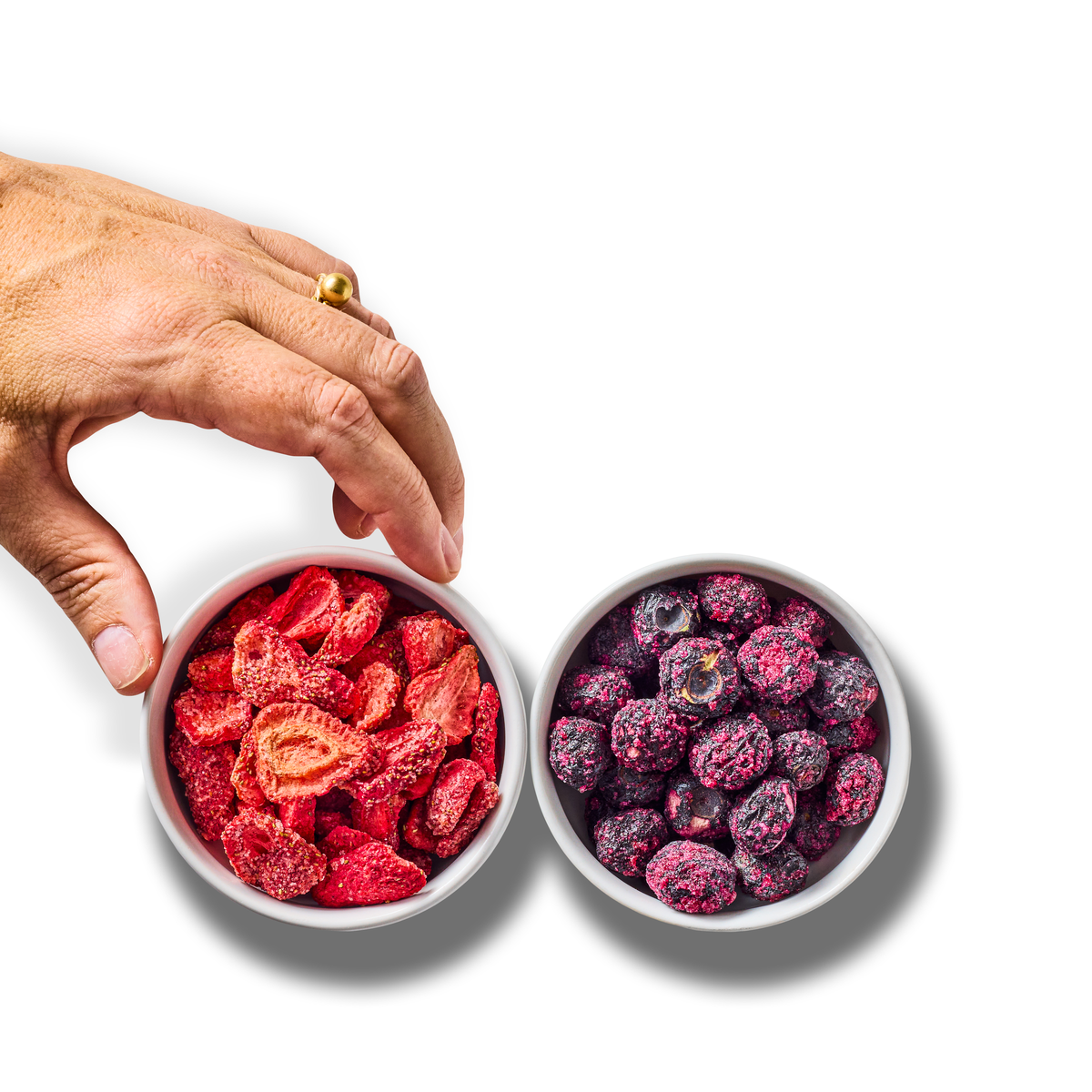 A hand reaches for a bowl of freeze-dried strawberries next to a bowl of freeze-dried blueberries, seen from above on white