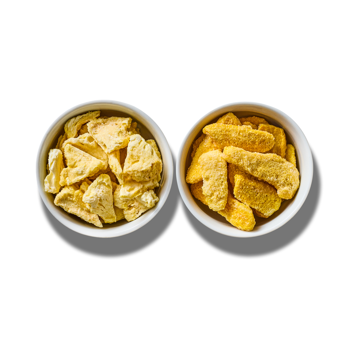 Tropical Blend two-pack: a white bowl of freeze-dried pineapple pieces beside a white bowl of freeze-dried mango pieces, seen from above on white