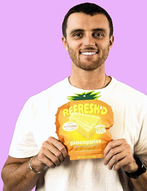 Man holding a pouch of REFRESH'D sour freeze-dried pineapple in both hands against a violet wall