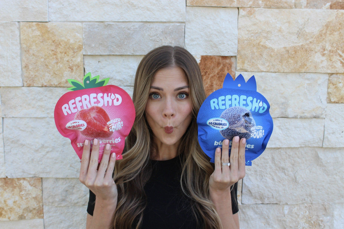 A woman making a sour face against a stone wall, holding REFRESH'D pouches of sour freeze-dried strawberries and blueberries