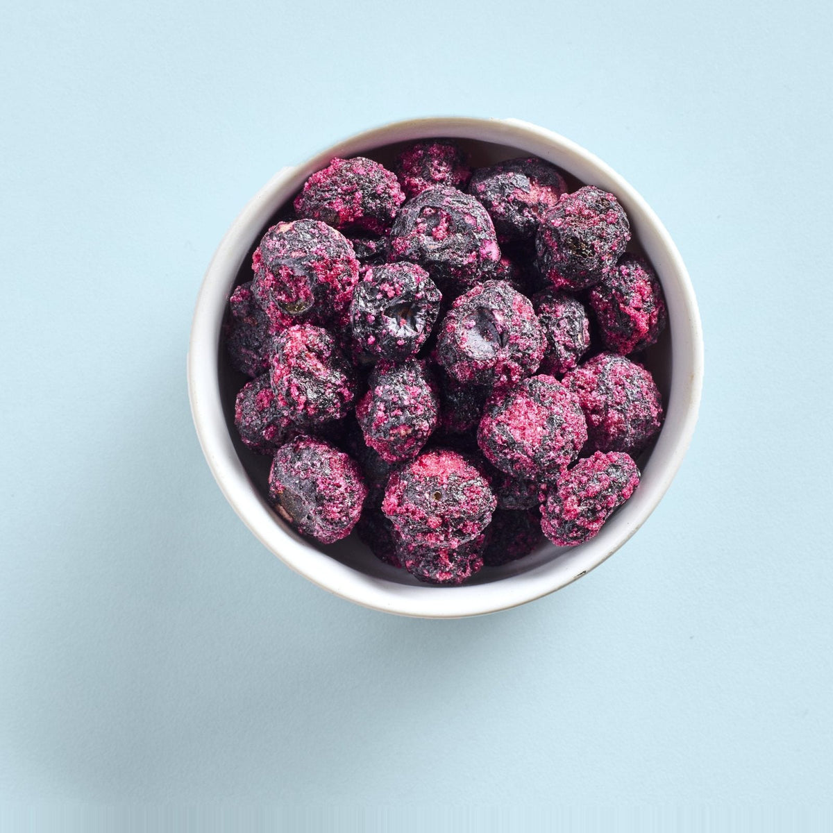 A white bowl of sour freeze-dried blueberries seen from above on a pale blue background