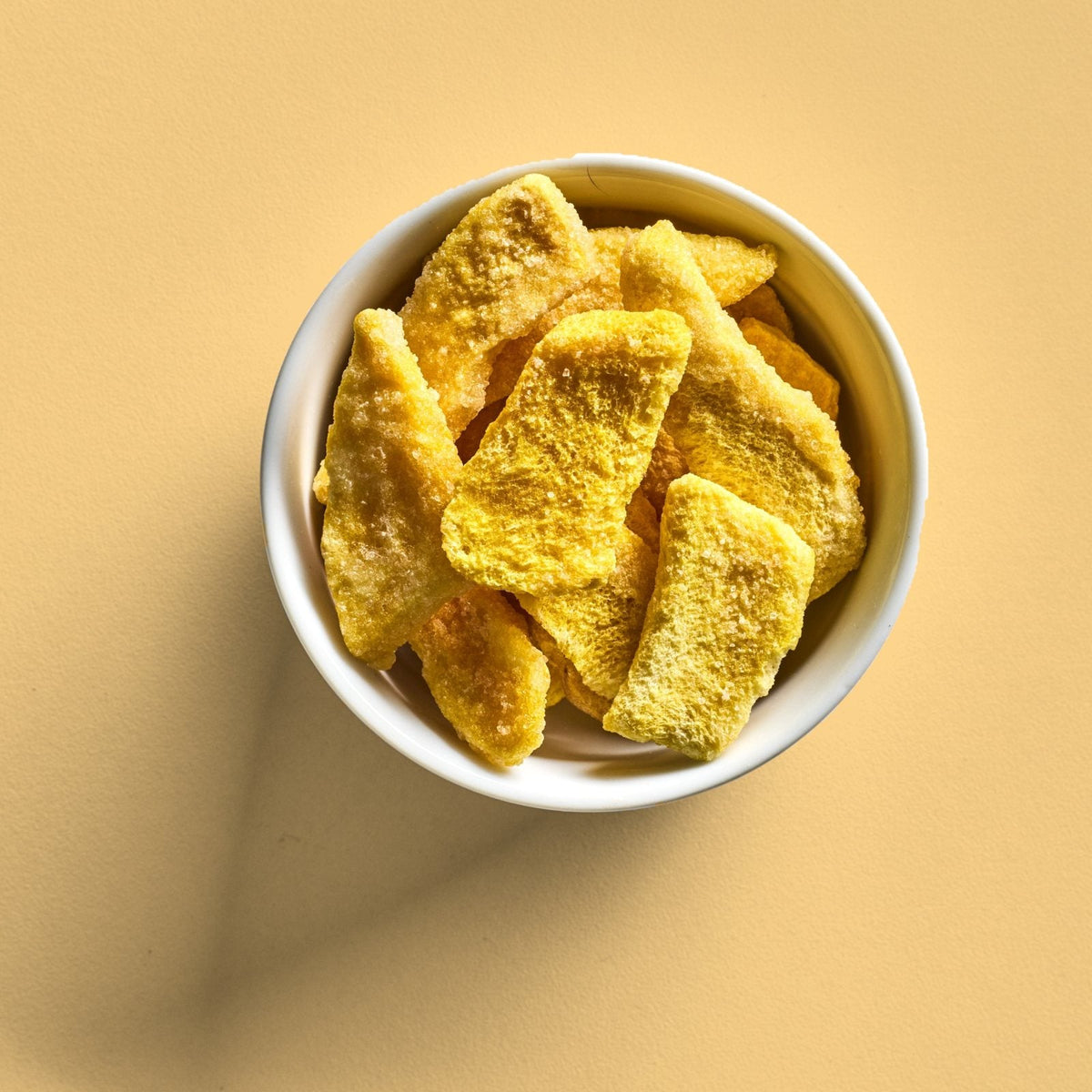 A white bowl of sour freeze-dried mangos seen from above on a yellow background