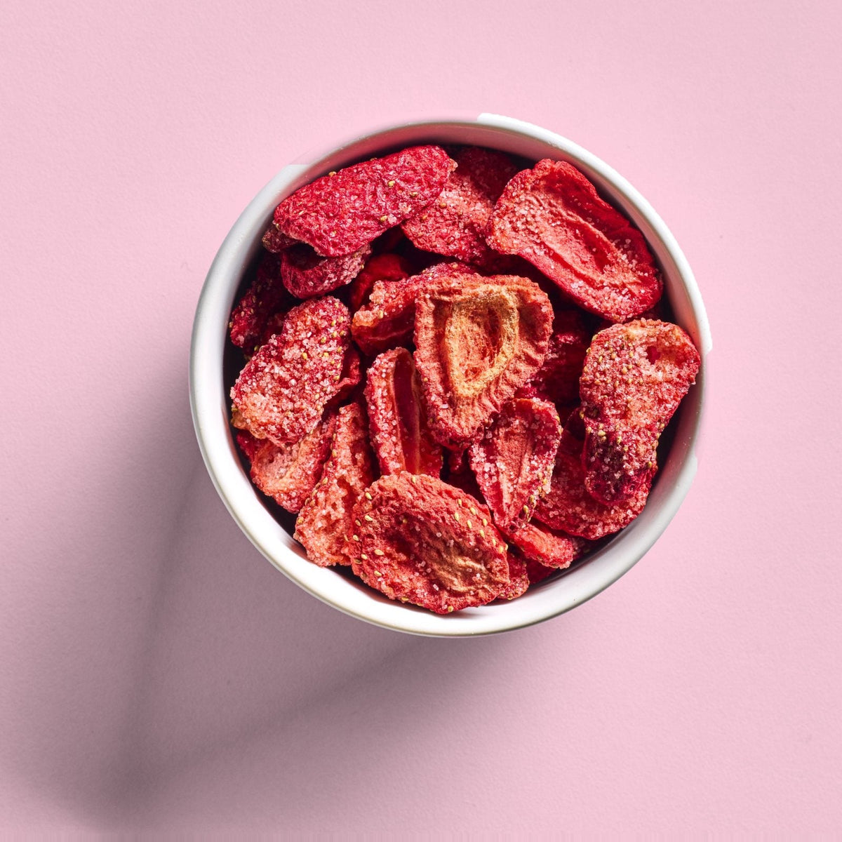 A white bowl of sour freeze-dried strawberries seen from above on a pink background
