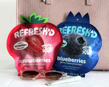 REFRESH'D strawberries and blueberries pouches laid flat on a white cloth beside a pale pink handbag and sunglasses