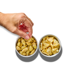 A hand reaches into a bowl of freeze-dried grapes beside a bowl of freeze-dried pineapple pieces, seen from above on white