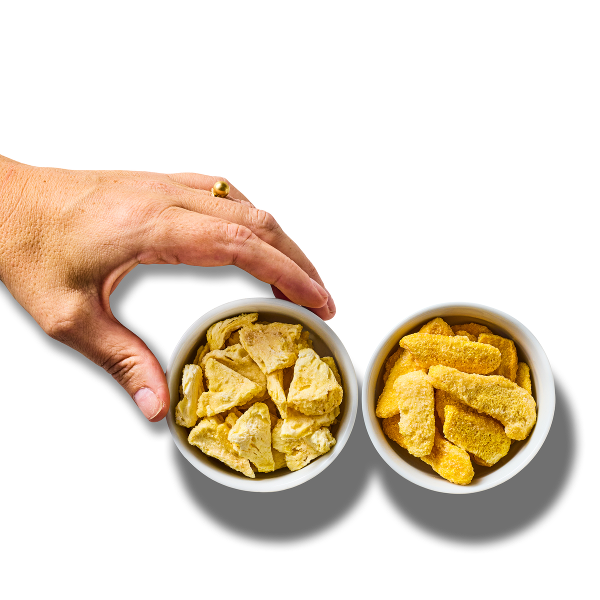 A hand reaches for a bowl of freeze-dried pineapple pieces next to a bowl of freeze-dried mango pieces, seen from above on white