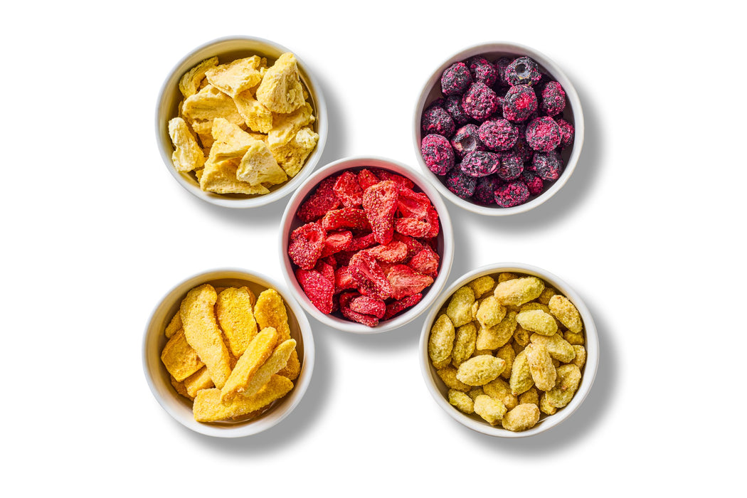 Five white bowls seen from above: sour freeze-dried pineapples, blueberries, strawberries, mangos and grapes