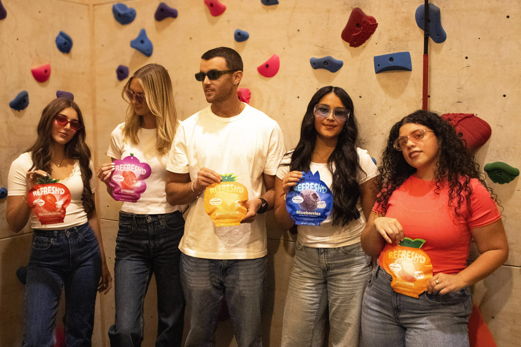 Five friends standing at an indoor climbing wall, each holding a REFRESH'D pouch of sour freeze-dried fruit