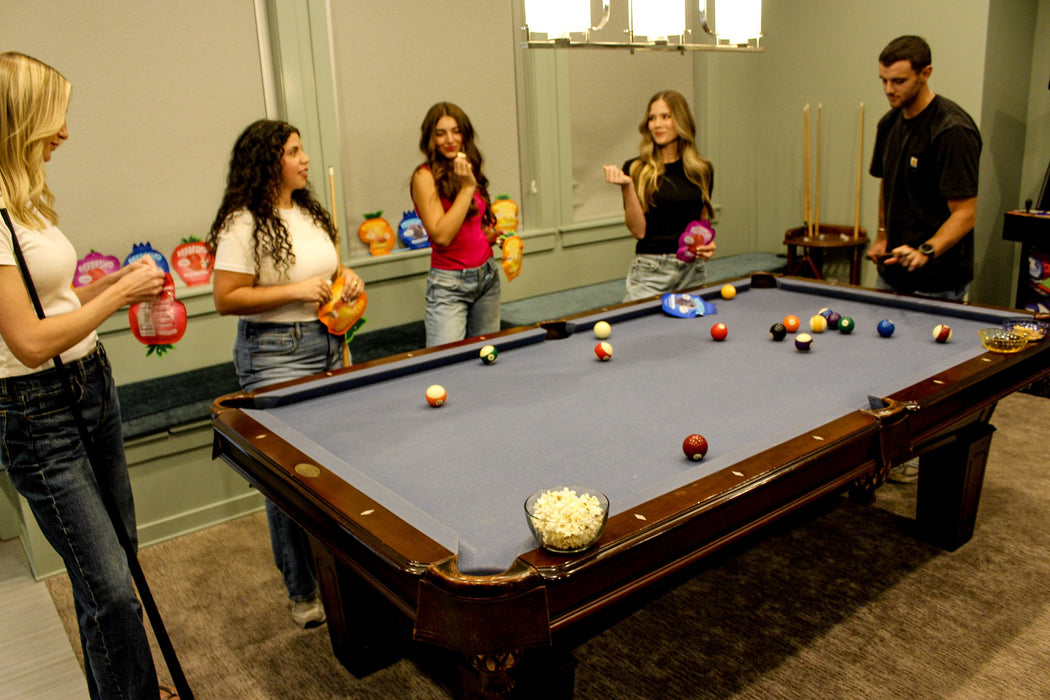 Friends around a pool table in a games room holding REFRESH'D pouches, a bowl of popcorn on the table