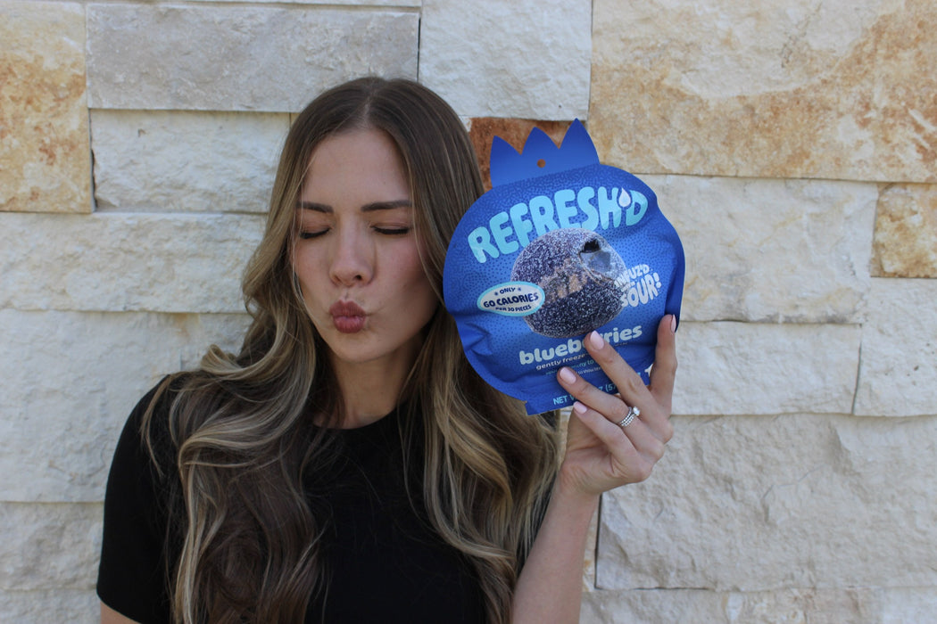 A woman with her eyes shut making a sour face, holding a REFRESH'D pouch of sour freeze-dried blueberries beside her cheek