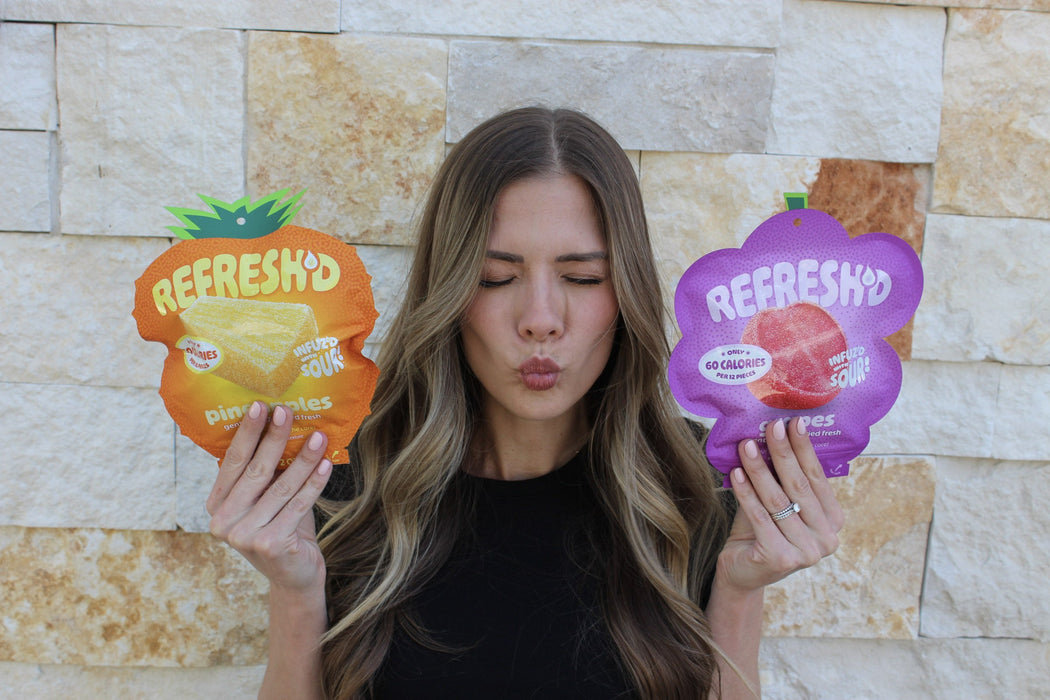 A woman with her eyes shut making a sour face, holding REFRESH'D pouches of sour freeze-dried pineapples and grapes against a stone wall