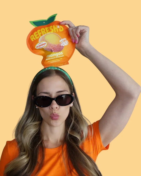 Woman lifting a pouch of REFRESH'D sour freeze-dried mango above her head against a peach wall