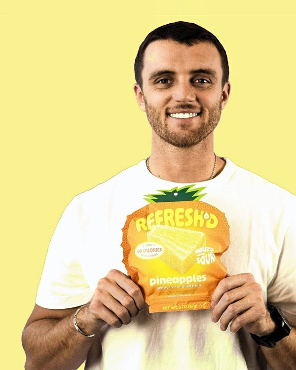 Man holding a pouch of REFRESH'D sour freeze-dried pineapple in both hands against a violet wall