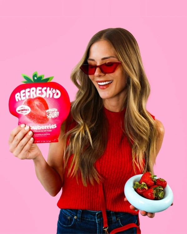 Woman in red holding a pouch of REFRESH'D sour freeze-dried strawberries and a bowl of fresh strawberries against a pink wall